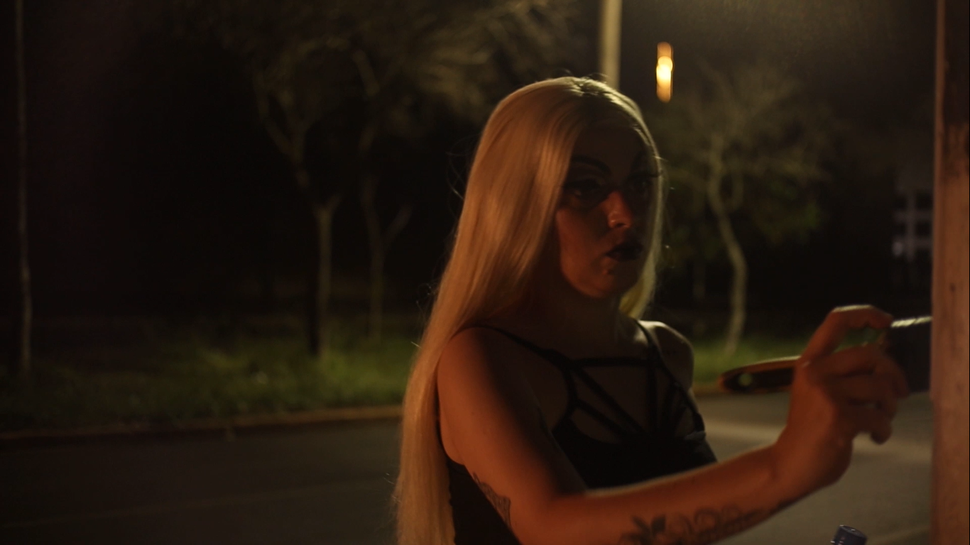 Pessoa com cabelo longo e loiro e maquiagem escura está ao ar livre à noite, segurando um objeto próximo a uma superfície de madeira com expressão concentrada. A iluminação fraca de cima projeta sombras, com árvores visíveis ao fundo.